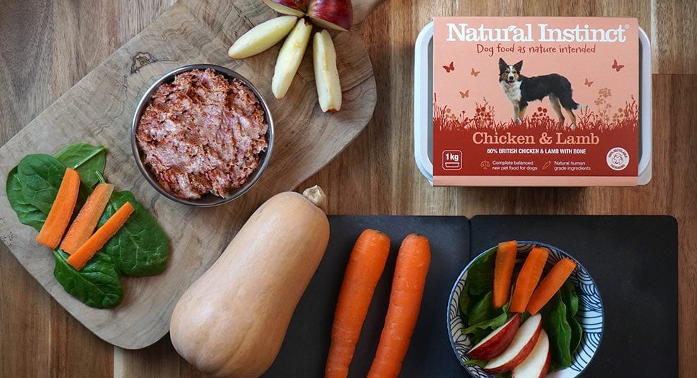 Natural Instinct Chicken and Lamb raw dog food packaging next to plated up food and ingredients displayed on slate and wooden boards