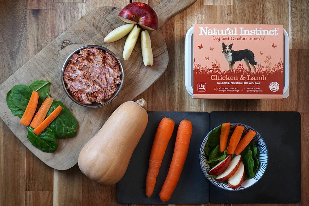 A wooden table with a cutting board holding spinach, carrot sticks, and a sliced apple; a bowl of minced dog food; a carton labeled Natural Instinct Chicken & Lamb; plus butternut squash, carrots, and a bowl of veggies.